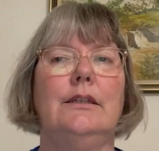 Angela – Leadership That Creates Results - Coaching College - Coaching College Close-up of a middle-aged woman with short gray hair and pink-rimmed glasses, looking at the camera.