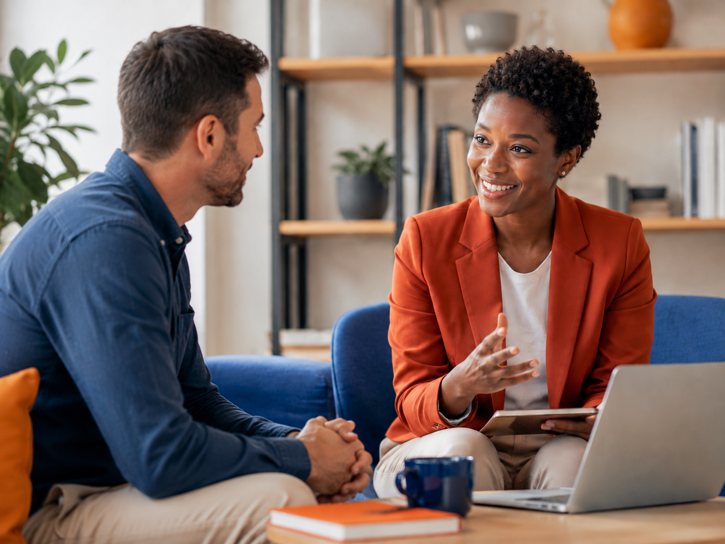 coaching 1 - Coaching College - Coaching College Two colleagues discuss results on a laptop/tablet in a casual office living room; woman in an orange blazer speaks with a smile.