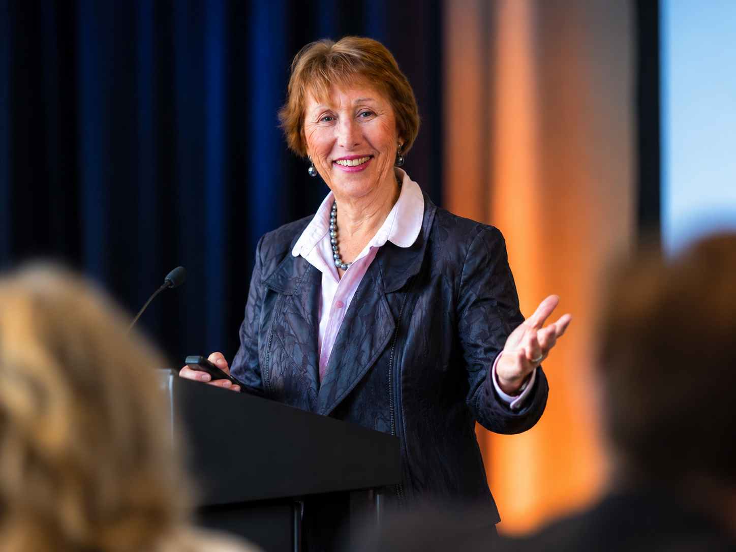 me speaking - Coaching College - Coaching College Smiling woman in a blazer giving a speech at a podium with a microphone, gesturing to the audience.